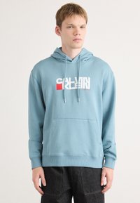 Svetlo modrá mikina s kapucňou a klokanie vrecko, s výrazným bielym a červeným grafickým logom na prednej strane a rebrovanými manžetami.