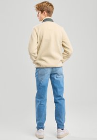 Giacca in pile crema, colletto alto con bordi verde scuro, abbinata a jeans azzurri chiari e sneakers bianche. Tessuto morbido, vestibilità casual.