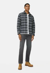 Veste à carreaux bleu, gris et noir, portée sur une chemise blanche. Un jean en denim gris et des bottes lacées beiges complètent la tenue.