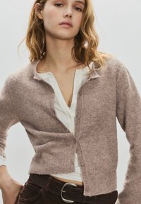 Tânără purtând un cardigan roz deschis cu nasturi peste un tricou alb henley și pantaloni maro închis cu o curea neagră.