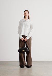 Biała bluzka w paski z kołnierzykiem, brązowe spodnie o szerokich nogawkach oraz czarna torebka z złotym zapięciem, stojące na szarej podłodze na tle jednolitej ściany.