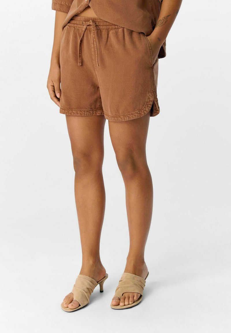Pantaloni corti marroni casual con vita elasticizzata e tasche laterali. Tessuto morbido con una superficie liscia. Abbinati a sandali beige con infradito.