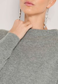 Nahaufnahme einer Person, die lange Kristalltropf-Ohrringe und einen grauen Strickpullover trägt, während die Hand ihren Hals berührt.