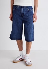 Tummansiniset denim-shortsit, joissa on rento malli ja polvipituinen leikkaus, etutaskut ja hienovarainen kuluminen. Yhdistetty valkoisten urheilukenkiä ja sukkia.