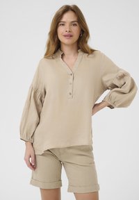 Beige vászon blúz gallérral, gombos kivágással és puffos ujjakkal; csíkos bézs sorttal viselve. Sima textúra, laza fazon.
