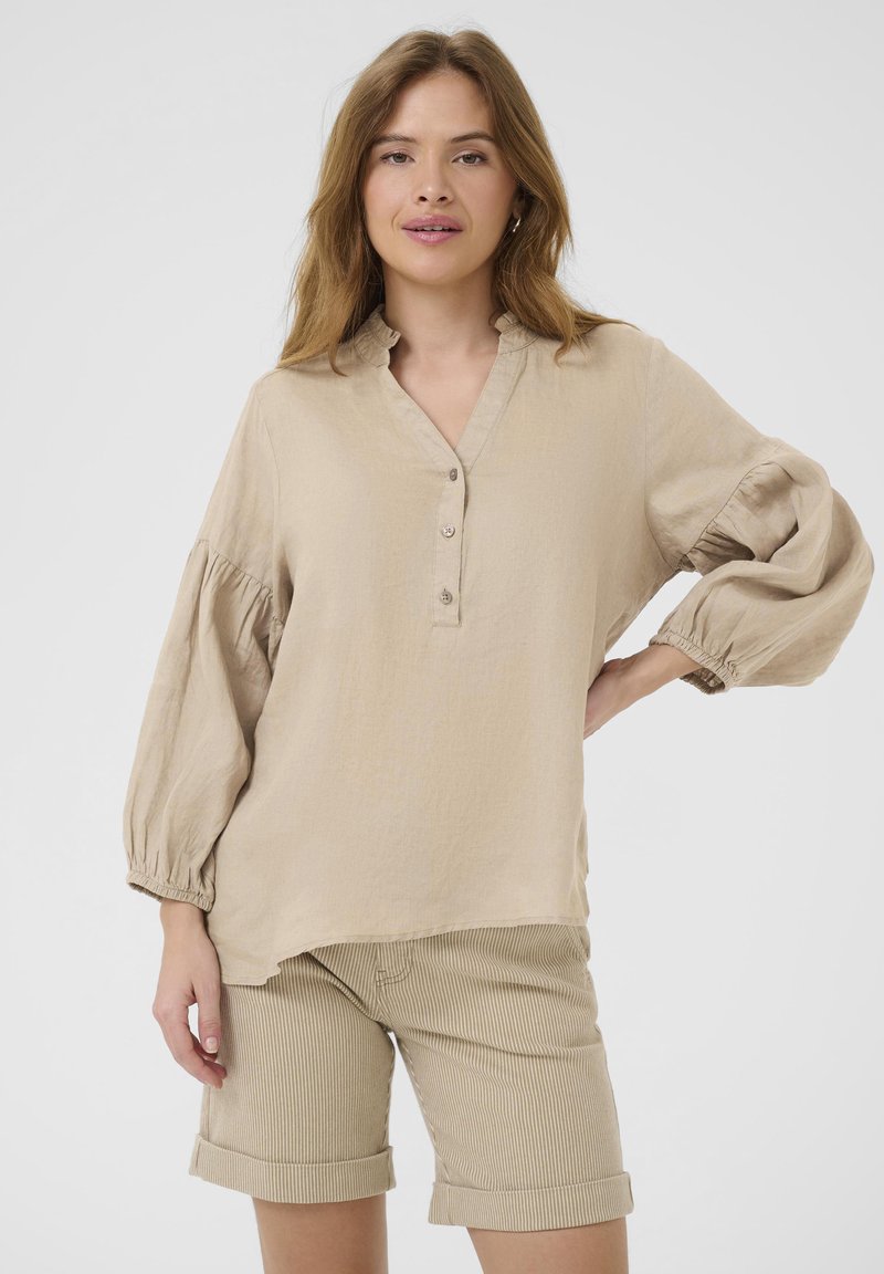 Blusa di lino beige con colletto, chiusura a bottoni e maniche a palloncino; indossata con pantaloni corti beige a righe. Texture liscia, vestibilità rilassata.