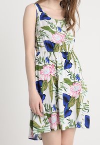 Květinové slunečné šaty s bílým pozadím, zdobené velkými růžovými pivoňkami a modrými květinami. Látka je lehká s uvolněným střihem.