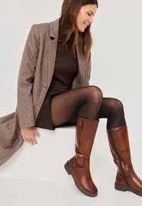 Stivali alti in pelle marrone con una texture liscia, punta rotonda, zip laterale e un dettaglio con fibbia sulla parte superiore, abbinati a un top e una gonna in maglia marrone.