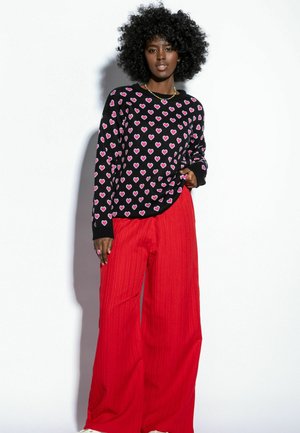 Czarny sweter w różowe wzory serc, zestawiony z szerokimi, czerwonymi spodniami z prążkowanej tkaniny. Ubranie ma długie rękawy i luźny krój.