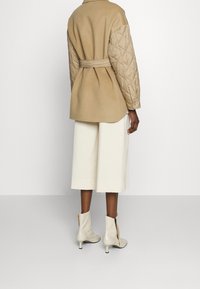 Veste matelassée couleur fauve avec une taille ceinturée, associée à des culottes crème et des bottines beiges. Matières lisses et texturées mises en avant.