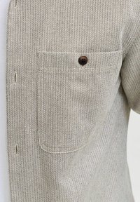 Lichtbeige overhemd met knopen, gemaakt van een gestructureerde stof, met een borstzak en een bruine plastic knoop.