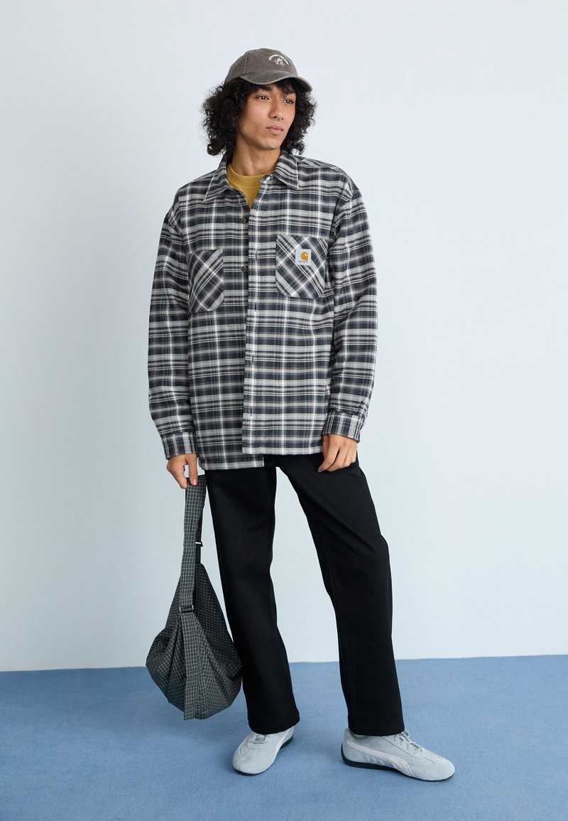 Geruite flanellen shirt in zwart en wit, met twee borstzakken. Zwarte broek en lichtgrijze sneakers. Grijze pet met logo. Grijze geruite tas.