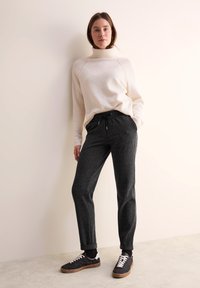 Sweatshirt beige clair à col montant, associé à un pantalon de jogging noir à motifs. Le modèle porte des baskets noires et blanches. Arrière-plan simple.