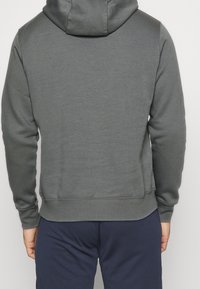 Sweat à capuche gris avec une capuche spacieuse, des poignets côtelés et un ourlet. Texture de tissu doux avec des coutures minimales. Vue de dos sans logos ni motifs visibles.