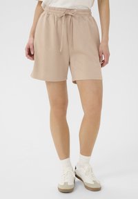 Pantaloni da ginnastica beige con vita elastica e coulisse, caratterizzati da una texture morbida e una vestibilità ampia. Abbinati a sneaker bianche.