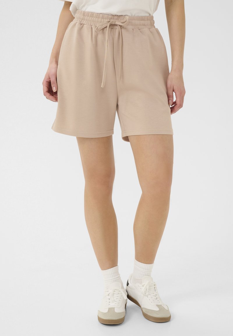 Pantaloni da ginnastica beige con vita elastica e coulisse, caratterizzati da una texture morbida e una vestibilità ampia. Abbinati a sneaker bianche.