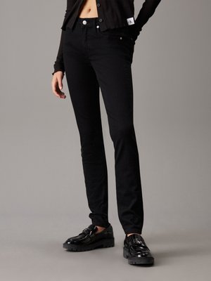 Jeans ajustados negros de mezclilla, con un diseño clásico de cinco bolsillos, cierre de botón, corte Slim y longitud hasta los tobillos. Llevados con zapatos brillantes.