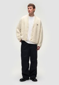 Cardigan bianco fluffoso con vestibilità morbida e scollo rotondo, abbinato a pantaloni cargo scuri e scarpe nere. Dotato di chiusura con bottoni e logo discreto.