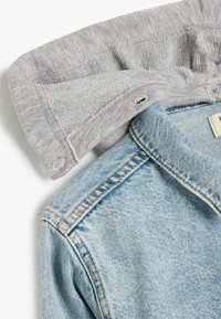 Collo di una felpa grigia chiara sovrapposto a una giacca di denim azzurro chiaro. Il denim presenta una texture sbiadita con cuciture visibili e un'etichetta.
