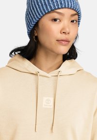 Beige huva sweatshirt med dragsnöre, som har en vit Timberland-logga på. Modellen bär en blå och vit randig stickad mössa.