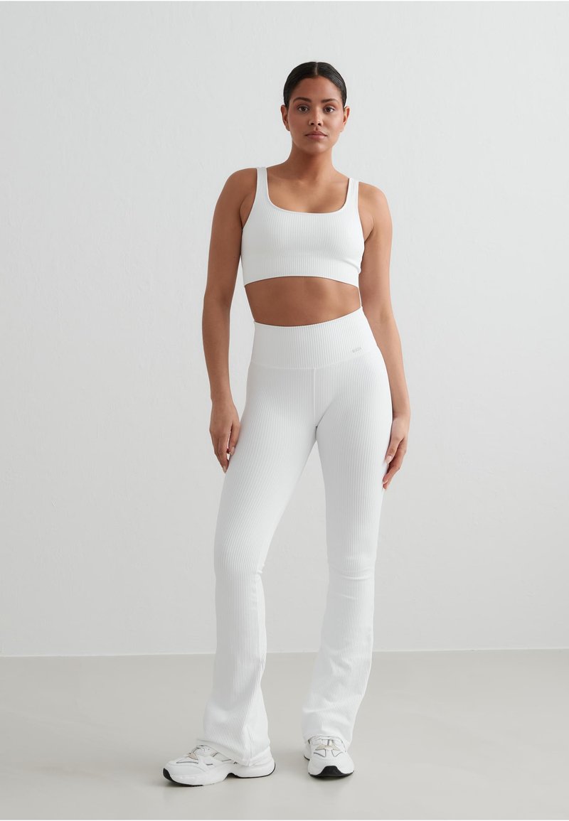 Hvit ribbestrikket aktivt antrekk sett som inkluderer en tettsittende crop top med brede stropper og høymidjede bukser med utsving, begge laget av et teksturert stoff.