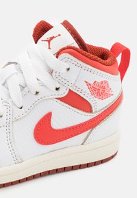 Sneaker alta bianca in pelle con accenti rossi, caratterizzata da un logo Swoosh Nike rosso, logo "Air Jordan" e lacci bianchi. Suola testurizzata.