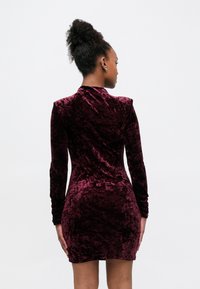 Robe bodycon en velours bordeaux avec manches longues, silhouette ajustée et tissu texturé. Présente un col montant et un détail froncé le long des manches.