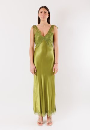 Donna in piedi che indossa un abito verde oliva senza maniche in raso con dettagli in pizzo, abbinato a tacchi bianchi aperti sulla punta, su sfondo bianco neutro.