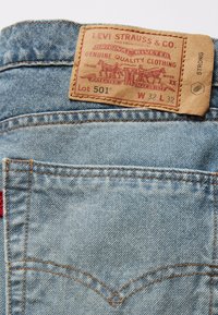 Jeans in denim azzurro chiaro con una toppa in pelle marrone. La toppa presenta scritte rosse e nere, un design con cavalli e dettagli rivettati.
