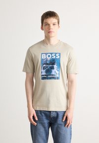 Tricou din bumbac bej cu un imprimeu grafic albastru, având textul "BOSS" și "PAN-AMERICAN" deasupra unei ilustrații a unui vehicul. Croială clasică.