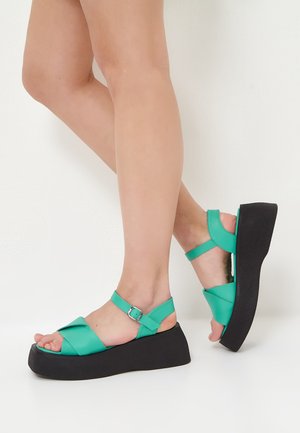 Sandali platform turchesi con suola nera, caratterizzati da un design a fascia incrociata e una fibbia alla caviglia regolabile. Texture piatta sulle fasce.
