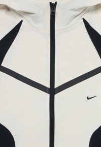 Gros plan sur une veste à capuche zippée noire et blanche, avec une bande diagonale noire et un petit logo Nike sur la poitrine.