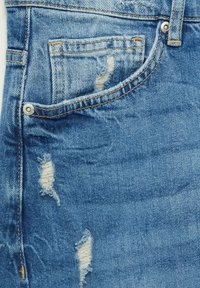 Primo piano di jeans in denim blu che mostrano la tasca anteriore con piccoli rivetti e dettagli di tessuto strappato vicino alla tasca e alla gamba dei pantaloni.