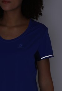 Blå kortärmad sporttröja i slät tyg, med reflekterande kant på ärmarna och en subtil logotyp på bröstet.