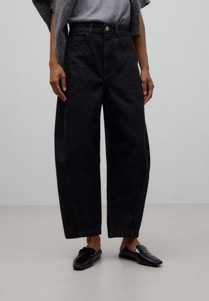 Person iført sorte jeans med høj talje og løs pasform, kombineret med sorte slip-on loafers, står op ad en ensfarvet grå baggrund.