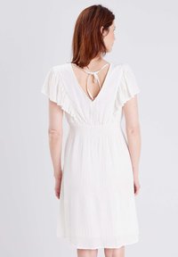 Weißes Kleid aus strukturiertem Stoff, kurze Rüschärmel und ein niedriger V-Ausschnitt, der mit einem Bündchen gebunden ist. Der Rock reicht bis über die Knie.