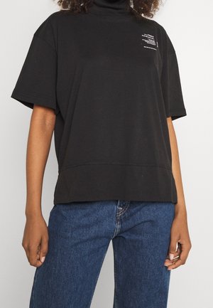 Personne portant un haut à col roulé à manches courtes noir ample avec un texte blanc sur la poitrine et un jean bleu en denim.