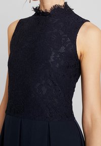 Mörkblå klänning med en spetskorsett med hög krage och blommönster, inramad av plisserad tyg i kjolen för en strukturerad silhuett.