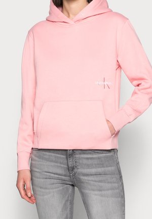 Osoba ubrana w jasnoróżową bluzę z kapturem Calvin Klein z kieszenią z przodu i szare jeansy, ręka w kieszeni, na jednolitym tle.