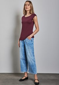 Bordová bezrukávová blůza s kulatým výstřihem, doplněná světle modrými širokými džíny. Modelka stojí před jednoduchým šedým pozadím.