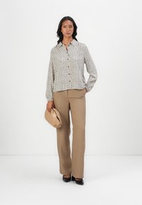 Hvit bluse med svarte prikkemønster, knapping foran, og lange puffede ermer; kombinert med brune rette bukser og en beige clutch-veske.