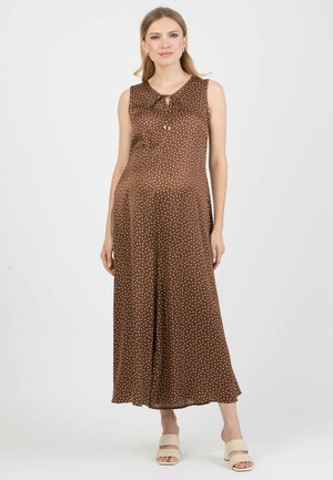 Žena v bezrukávých hnědých dlouhých šatech s bílými puntíky a zavazovacím detailem u výstřihu, doplněných krémovými sandálkami na podpatku.