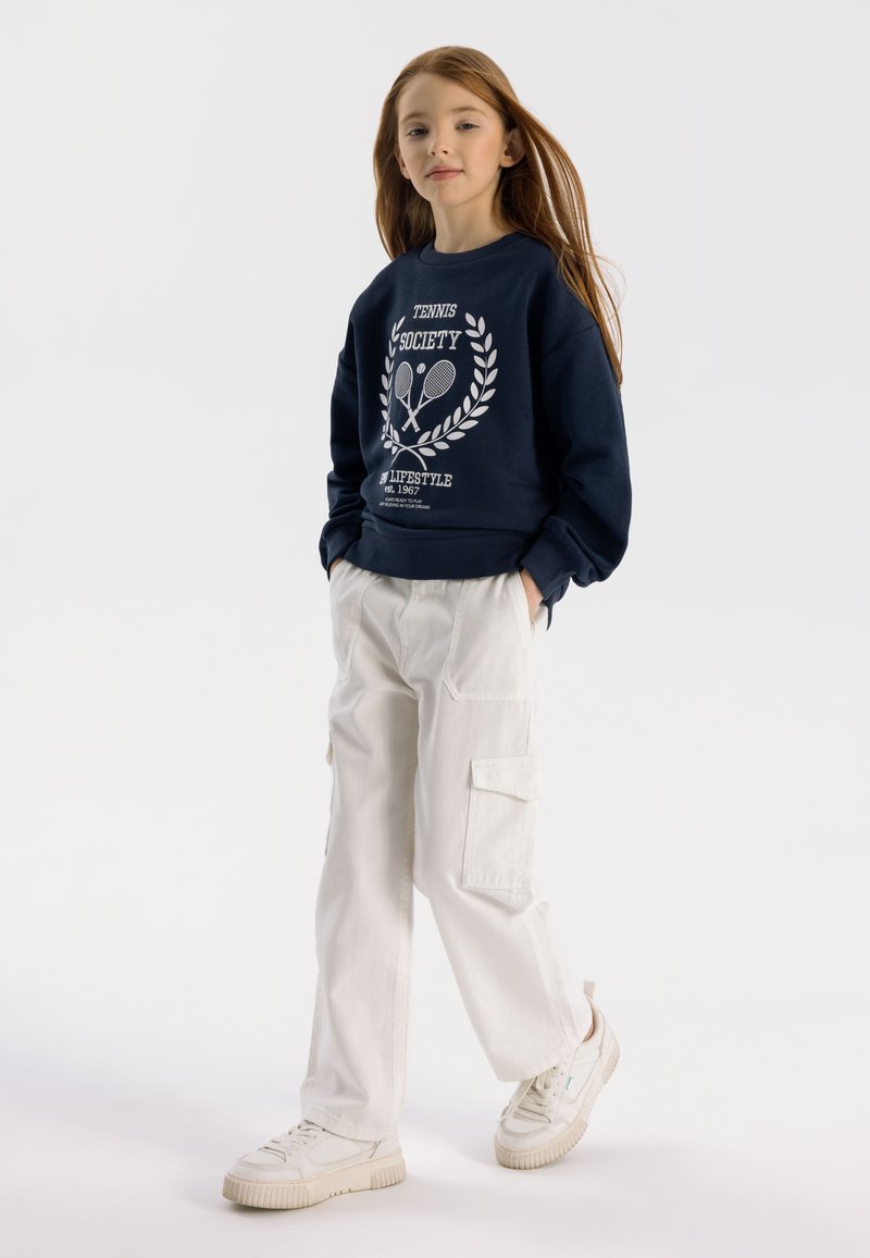 Sudadera de color marino con un gráfico blanco que dice "Tennis Society", combinada con pantalones cargo crema y zapatillas blancas. Textura suave, corte holgado.