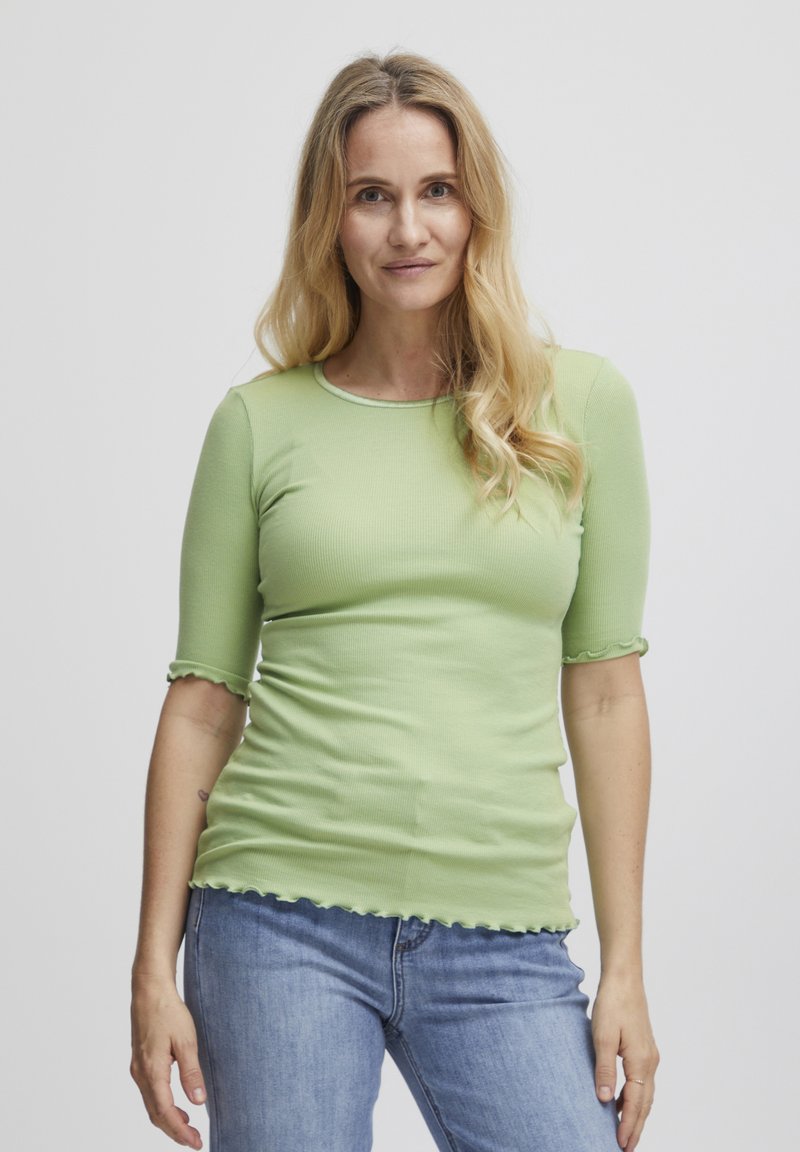 Top ribbed de culoare verde deschis, cu mâneci 3/4, având margini ondulate, asociat cu blugi albaștri. Textură netedă, design ajustat.