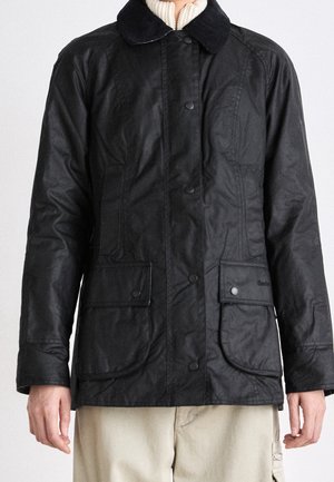 Schwarze gewachste Jacke mit Knopflaschen, aufgesetzten Taschen und Kragen, getragen über einem cremefarbenen Strickpullover mit Reißverschluss und beiger Hose.