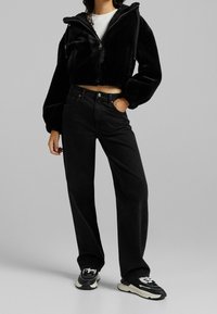 Giacca corta nera in pelliccia sintetica con cappuccio e vita elasticizzata. Abbinata a jeans neri a vita alta e sneakers nere con dettagli bianchi.