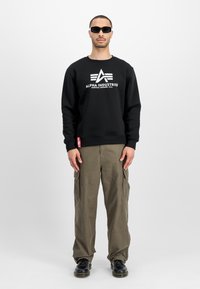 Uomo che indossa una felpa nera Alpha Industries, pantaloni cargo oliva, scarpe nere e occhiali da sole neri, in piedi su uno sfondo bianco.