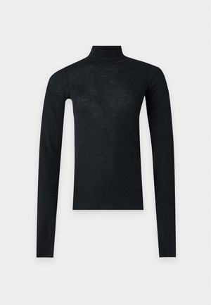 Femme debout portant un col roulé noir, un jean bleu clair taille haute et des bottes à talons pointus noirs sur un fond uni.