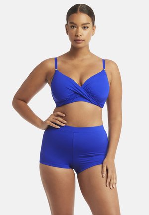 Vrouw staand met hand op heup, gekleed in een koningsblauwe sport-bh met kruisbanden aan de voorkant en bijpassende hoge taille shorts.