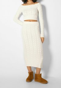 Knit two-piece set in off-white, featuring a cropped, off-shoulder top and a long, ribbed skirt with a textured pattern. Worn with tan boots.